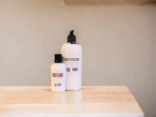 A small bottle of lotion, next to a large bottle of lotion, sitting on a light wood surface, with a tan background. 