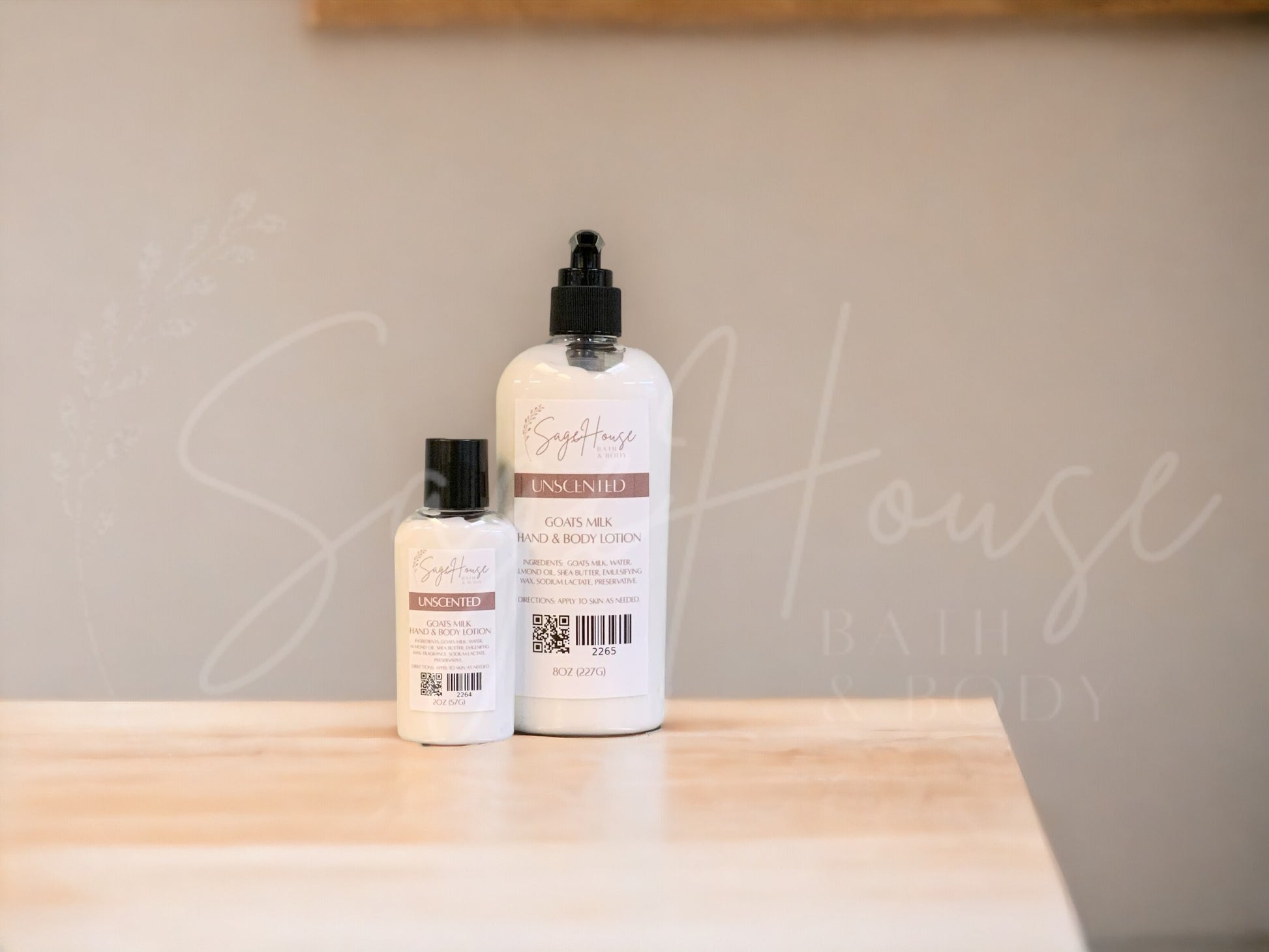 A small bottle of lotion next to a large bottle of lotion, sitting on a light wood surface, with a tan background. Has SageHouse logo inlayed