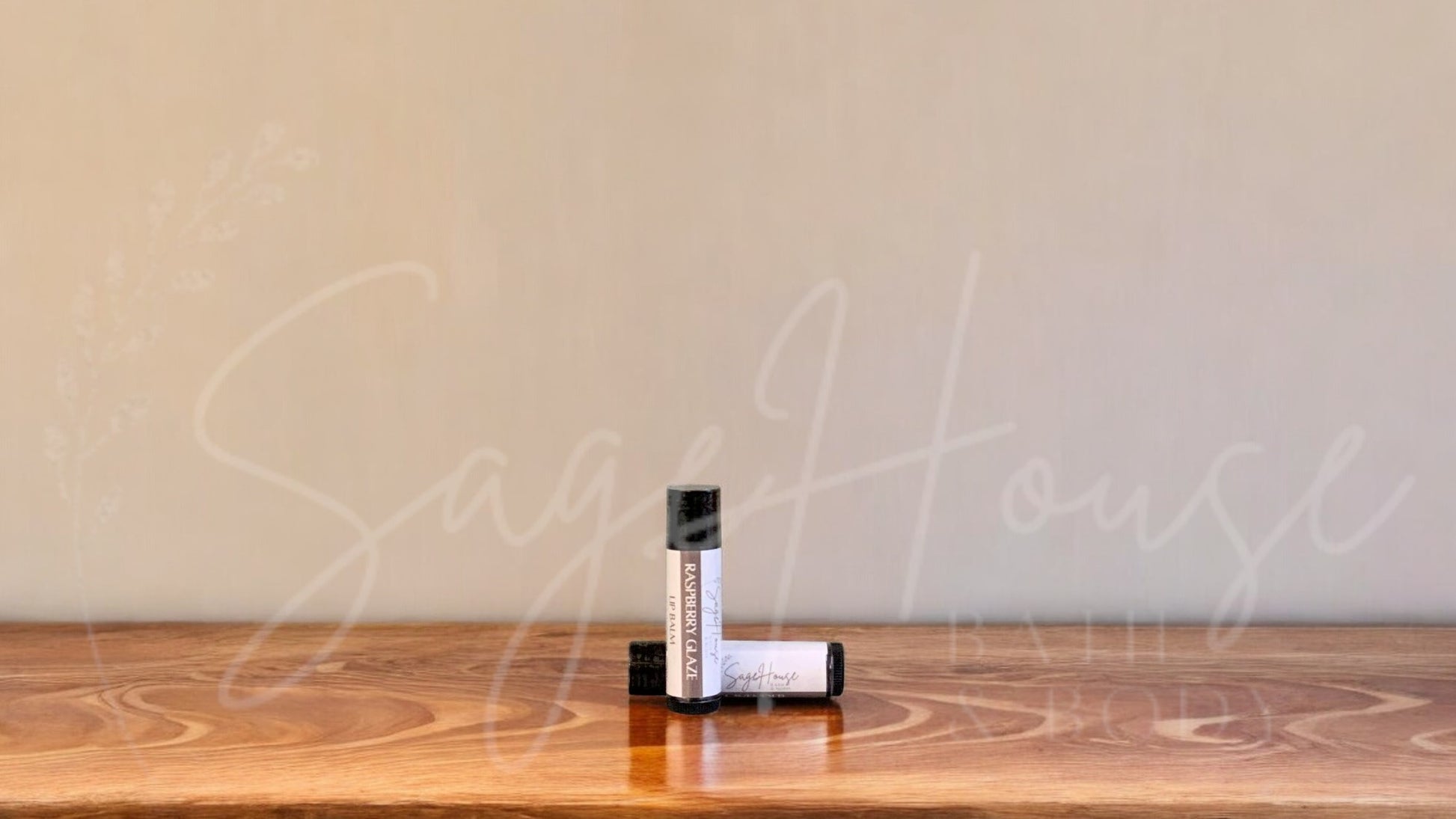 A tube of raspberry glaze lip balm placed upright with a tube lying behind it, on a wooden surface.