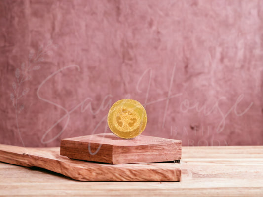 A yellow bar of raspberry lemonade loofah soap resting on a wooden soap dish, with a pink background.