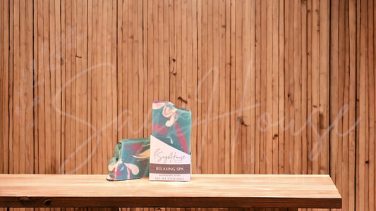 A teal colored bar of soap with accents of mauve and white, in a white labeled box, on a wood surface.