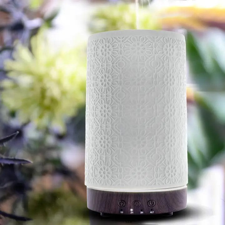 A white ceramic diffuser with a geometric floral pattern, featuring a top button and a base compartment, placed near flowers.