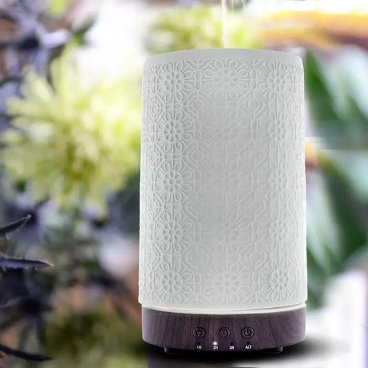 A white ceramic diffuser with a geometric floral pattern, featuring a top button and a base compartment, placed near flowers.