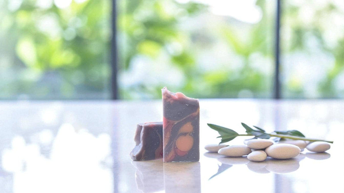 A purple and pink soap bar on a white surface with stones and greenery next to it, as well as greenery in the background