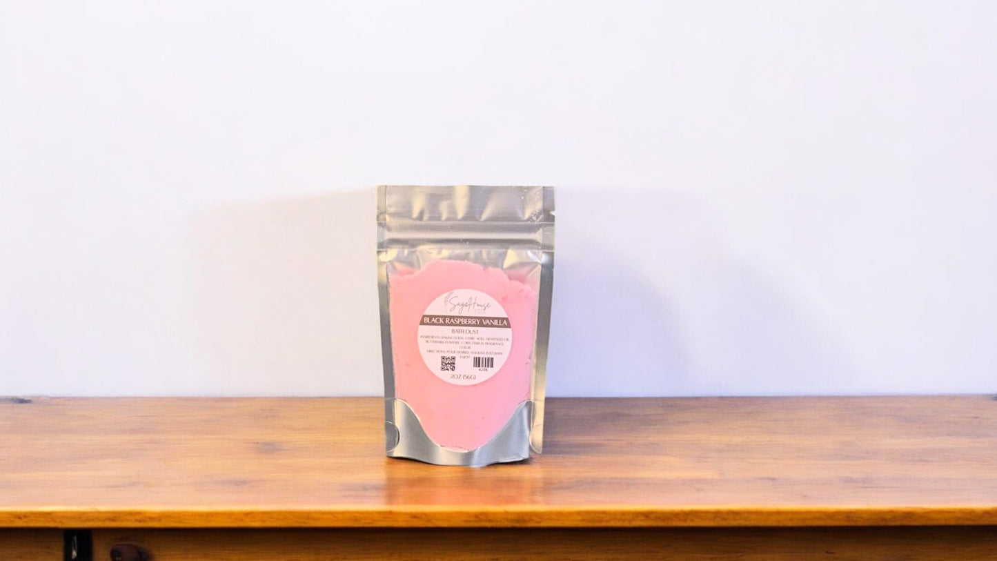 A pink-colored bath bomb in clear packaging, labeled 'black raspberry vanilla'. The bath bomb is placed on a wooden surface.