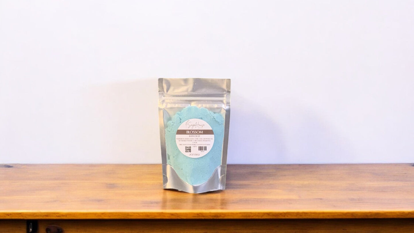 A teal-colored bath bomb in clear packaging, labeled 'blossom'. The bath bomb is placed on a wooden surface.