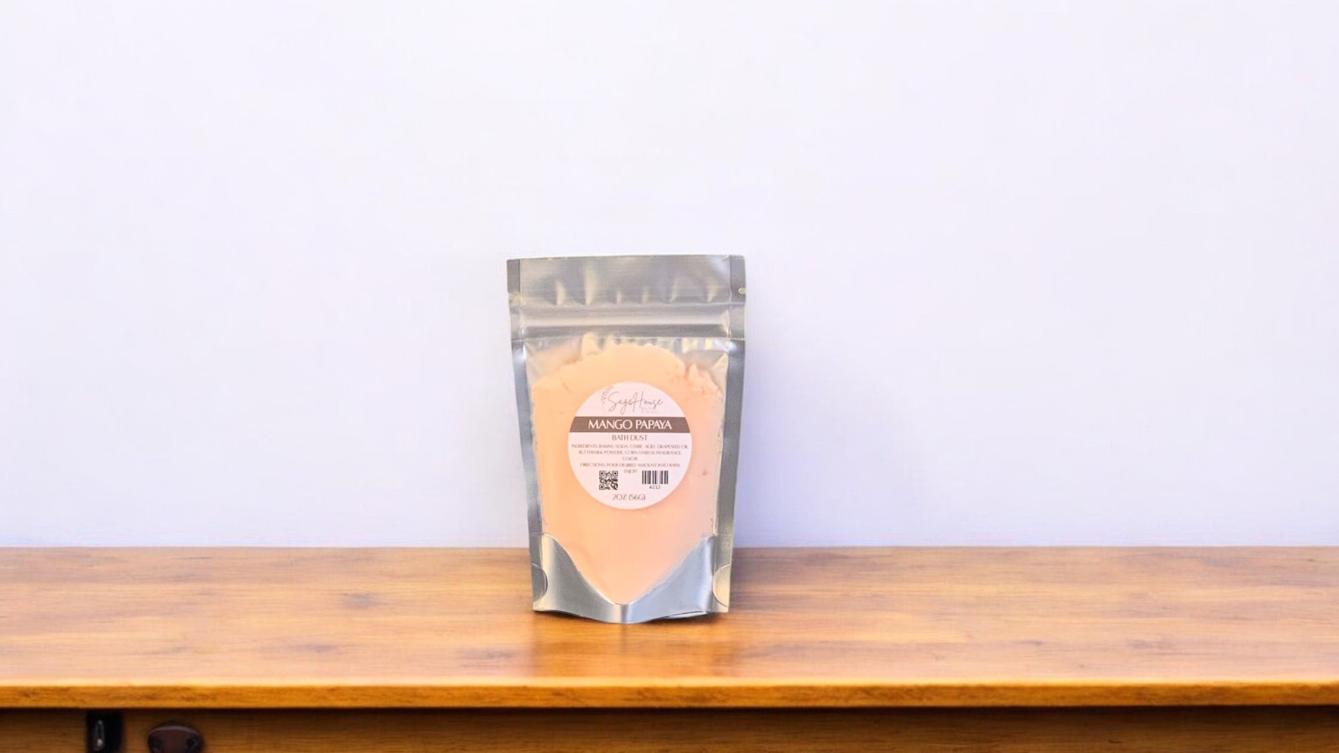 A peach-colored bath bomb in clear packaging, labeled 'mango papaya'. The bath bomb is placed on a wooden surface.