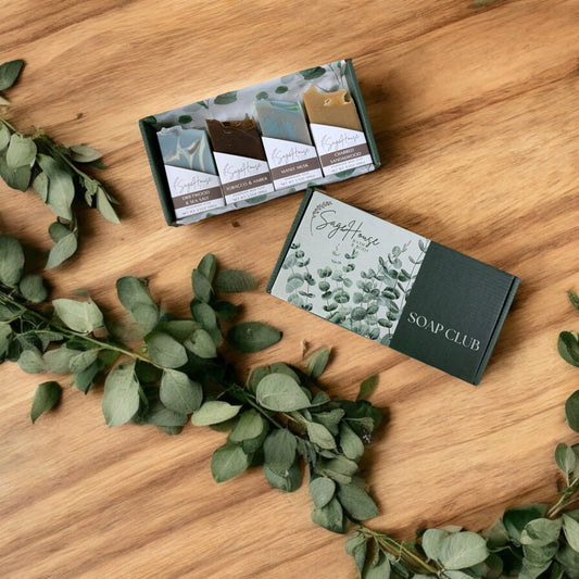 Four packaged soap bars inside of a box, arranged on a wooden surface with leaves scattered around. Front of box is labeled with the Soap Club branding.