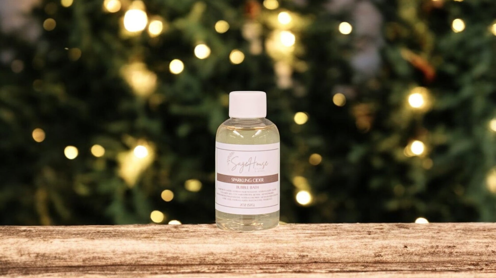 A small bottle of bubble bath, with white lid. On a wood top with greenery in the background.