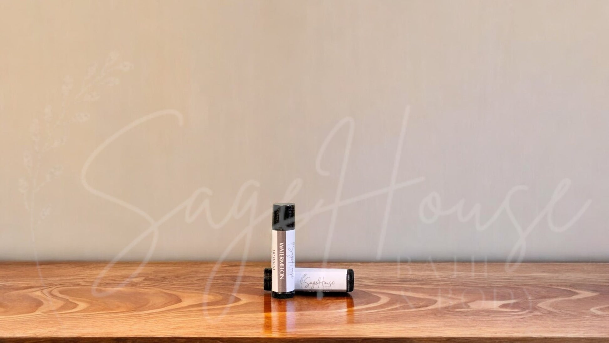 A tube of watermelon lip balm placed upright with a tube lying behind it, on a wooden surface.