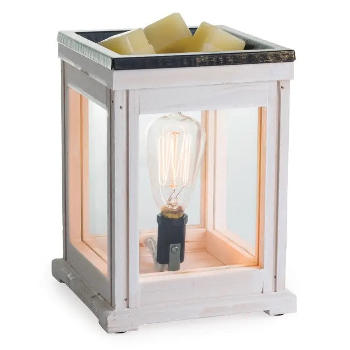 A weathered wood warmer with a clear glass enclosure and a halogen bulb, heating a dish with wax melts inside, creating a warm glow.