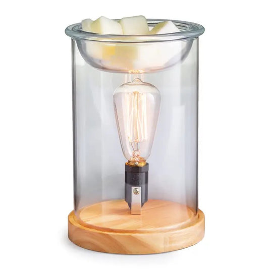A clear glass illumination fragrance warmer with a vintage bulb and metallic finishes, placed on a wooden base.